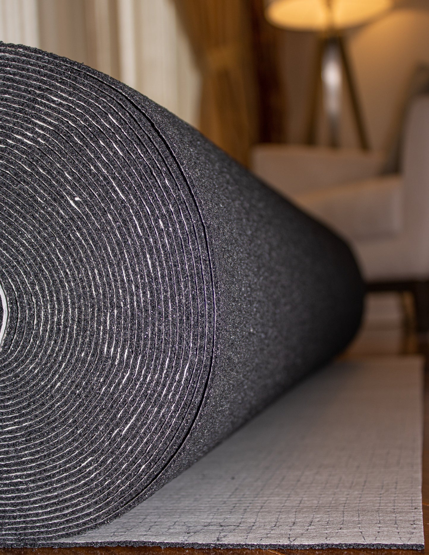 Close-up of a rolled dark gray Dura-Son™ 3.5 mm yoga mat by Dura Undercushions on a light gray carpet in a cozy room with a lamp and a couch in the background, offering sound reduction akin to an acoustic underlayment.