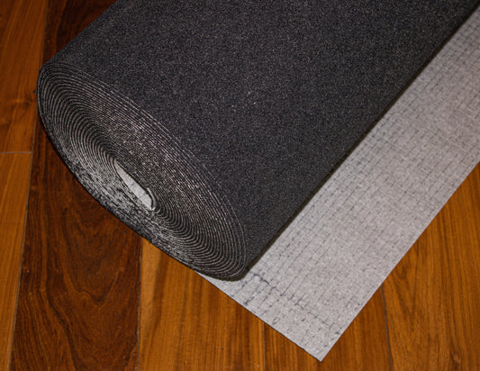 A large roll of Dura Undercushions' Dura-Son™ 3.5 mm black acoustic underlayment is partially unrolled on a wooden floor, revealing the underside with its grid pattern designed for sound reduction.