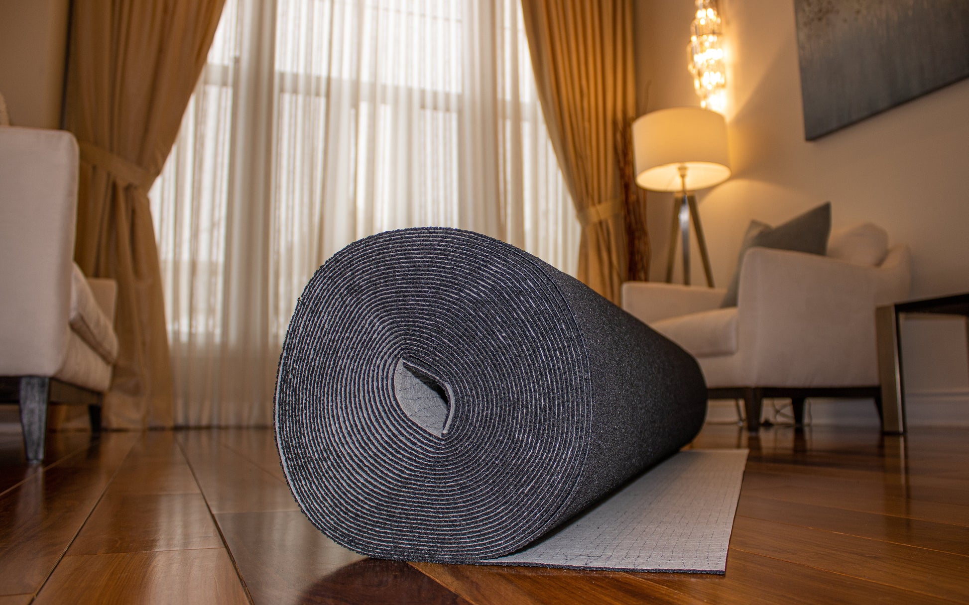 A rolled-up black yoga mat on a gray Dura-Son™ 3.5 mm by Dura Undercushions placed on a wooden floor in a cozy living room with a floor lamp, chairs, and curtains in the background.