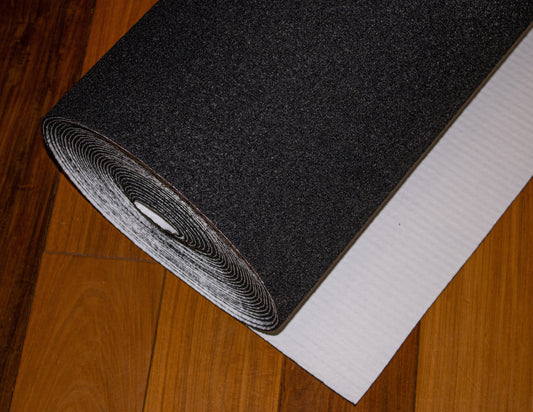 A roll of Dura Undercushions Dura-Son MB™ 3.5 mm black felt material partially unrolled onto a wooden floor, with a white backing visible underneath, designed to enhance acoustic performance.