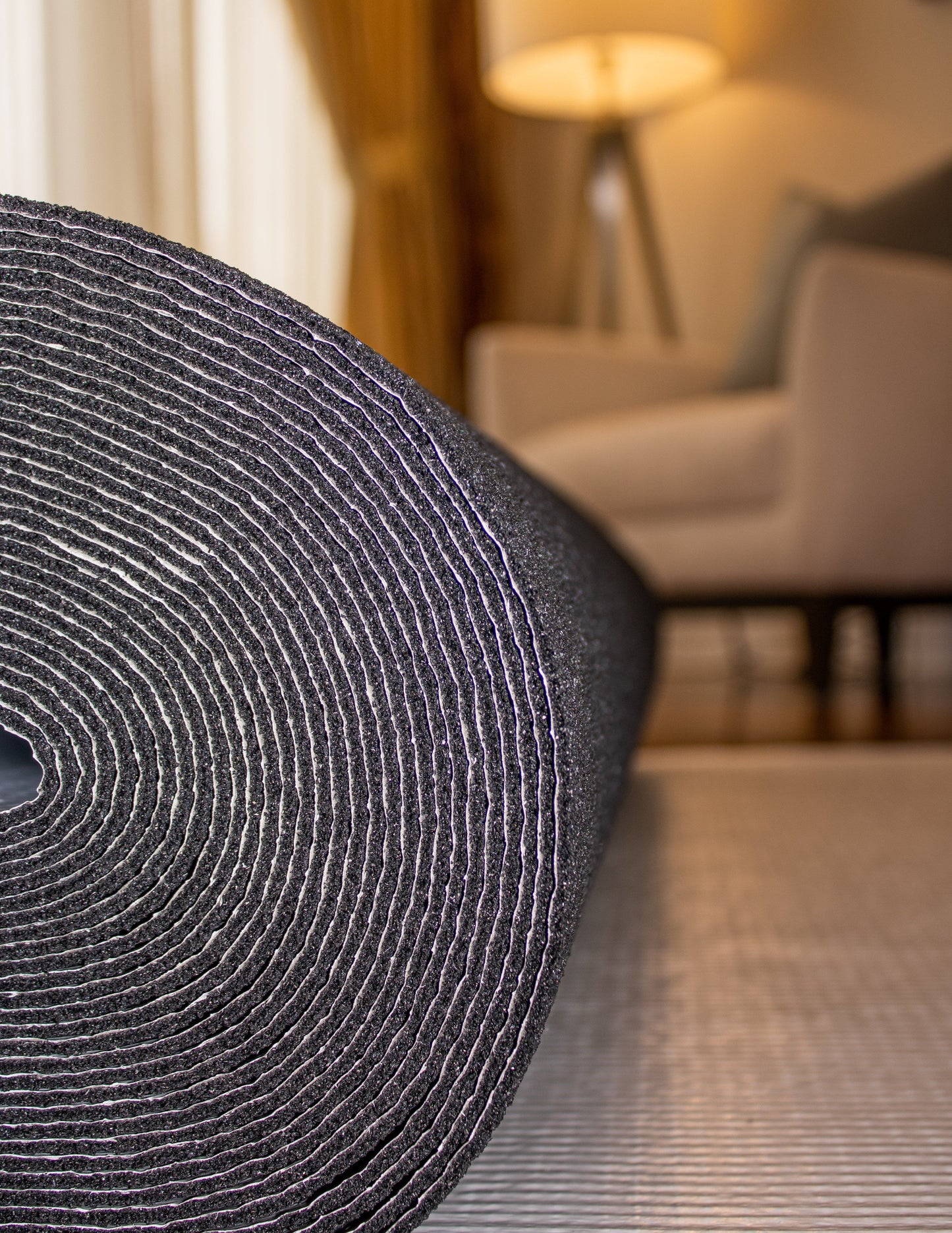 A close-up of a rolled-up black yoga mat on the floor of a living room with a beige armchair, table lamp, and brown curtains in the background showcases how the Dura-Son MB™ 3.5 mm by Dura Undercushions can complement any serene space.