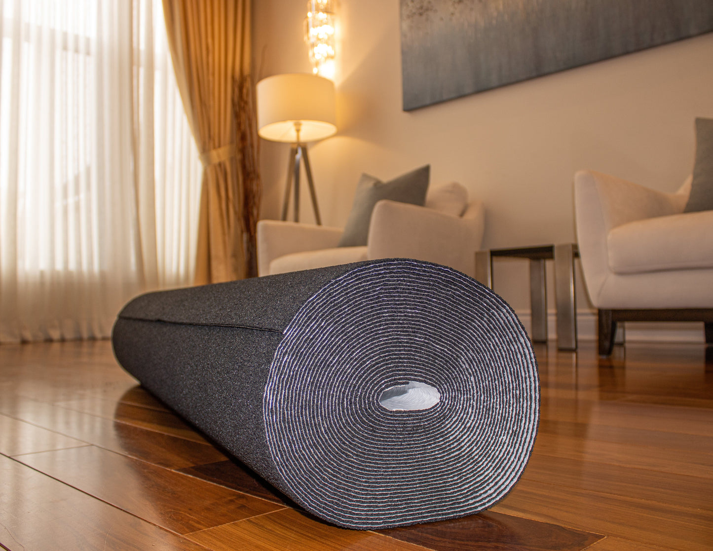 A rolled-up black Dura-Son MB™ 3.5 mm yoga mat by Dura Undercushions is placed on a wooden floor in a well-lit living room with a lamp, armchairs, and a painting in the background, offering an inviting space with enhanced acoustic performance.