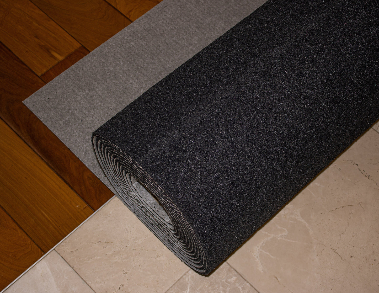 Close-up image of a roll of dark grey felt, Duracoustic™ 8 mm by Dura Undercushions, known for its superior impact sound reduction, lying on a grey and beige tiled surface next to a wooden floor.