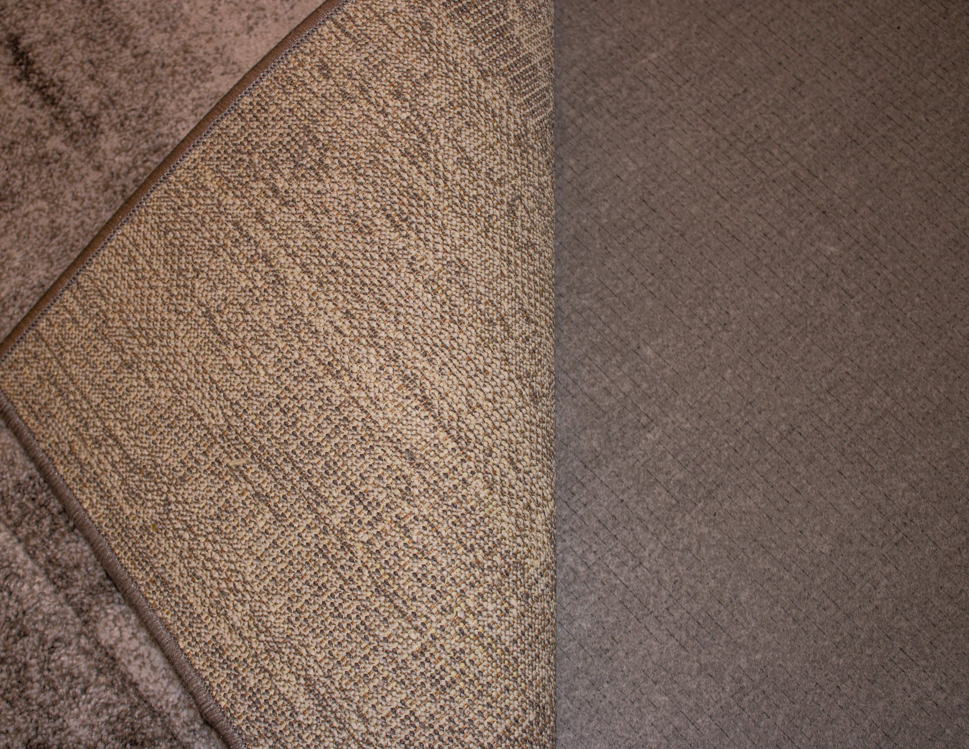 A close-up image shows a corner of a beige woven rug turned up to reveal a dark grey textured surface underneath, which enhances the Duracushion™ 6 mm by Dura Undercushions' acoustic performance.