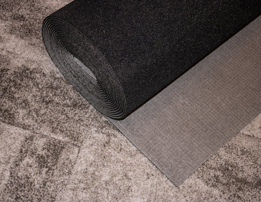 A rolled black and gray foam mat, partially unrolled on a textured, mottled surface, showcases its superior acoustic performance. Ideal as a carpet underlay, the Duracushion™ 6 mm by Dura Undercushions provides added comfort and enhances sound insulation.