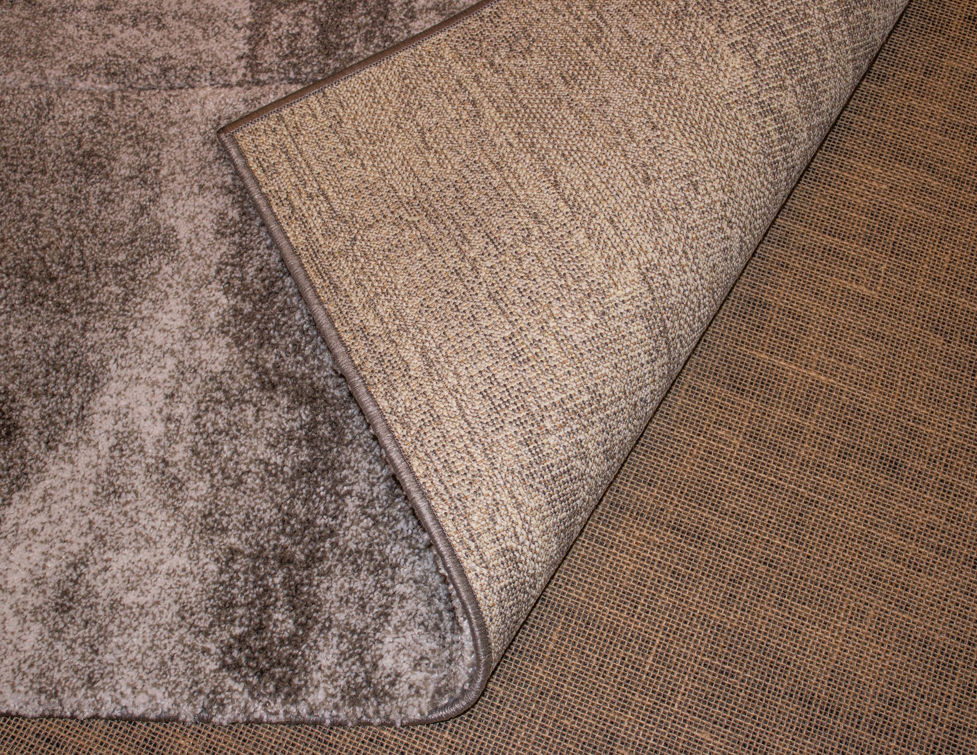 A corner of a textured gray rug is flipped over on a brown woven carpet, revealing its light underside and suggesting Dura Undercushions' Duralux™ Comfort 7 mm for high-end rug protection and improved acoustic performance.