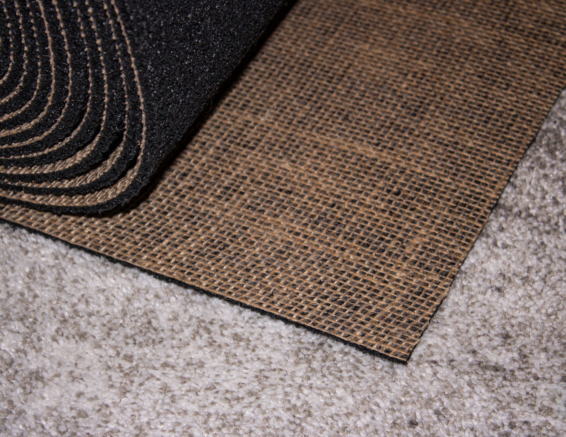 A close-up of the Duralux™ Comfort 7 mm by Dura Undercushions, a black rubber mat for premium rug protection, partially overlaps textured brown fabric on a gray carpeted floor.