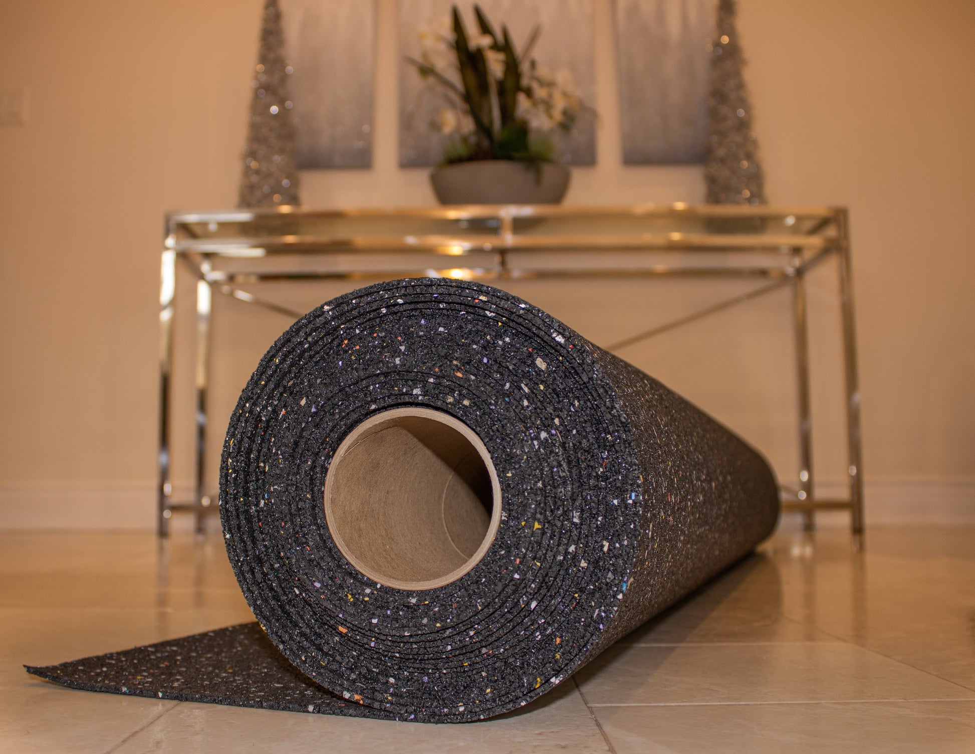 A roll of Duraquiet™ 3 mm black underlayment with colorful specks from Dura Undercushions is placed on a light-colored tile floor. A metallic table and small decorative trees are in the background.