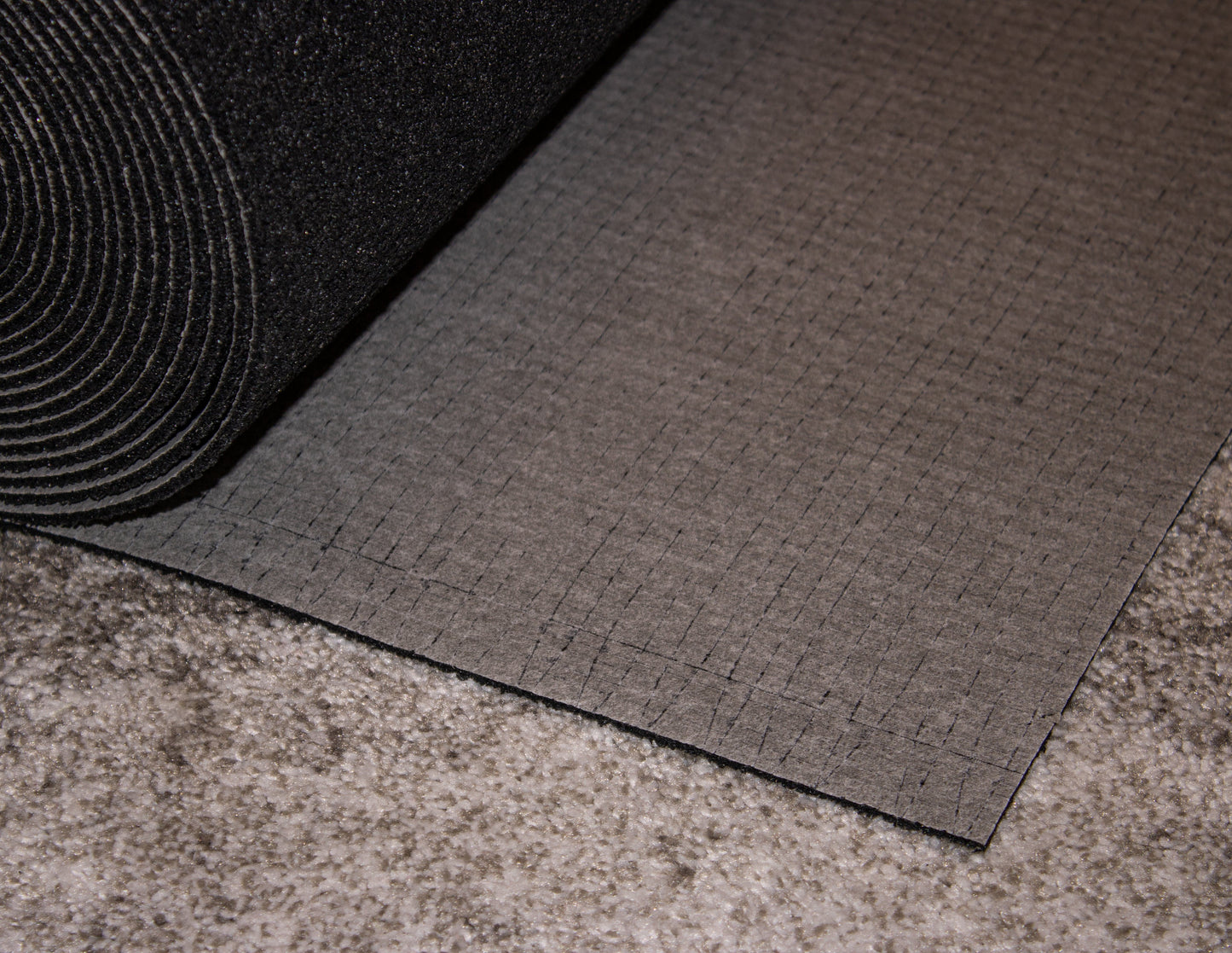 Close-up of a partially unrolled Protector™ 5 mm black foam mat by Dura Undercushions on a gray carpeted floor. The foam mat, designed for impact sound reduction, has a textured surface with a grid pattern.