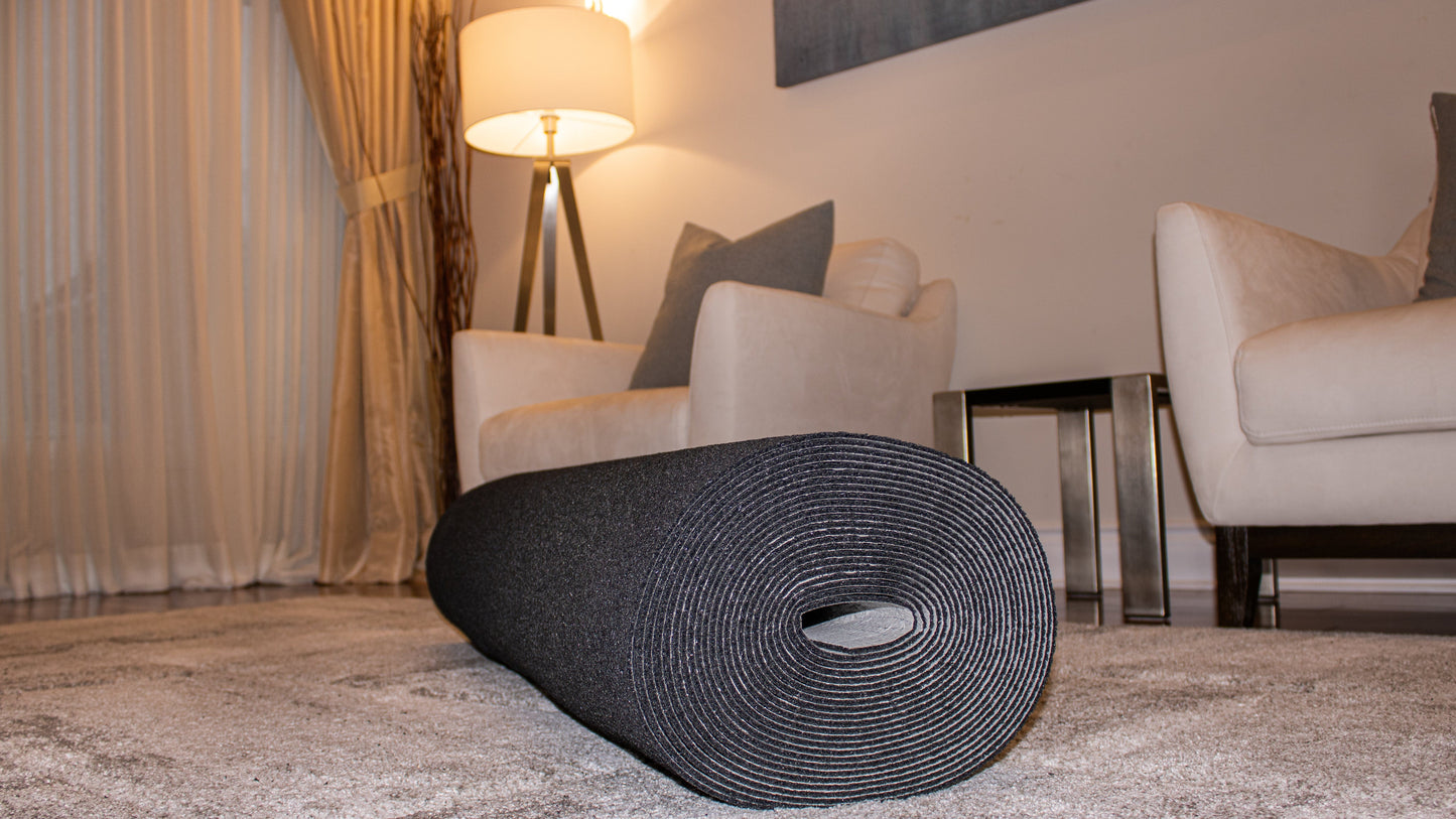 A rolled-up black yoga mat lies on a Super Dura™ 10 mm by Dura Undercushions in a living room with white armchairs, a side table, and a lit floor lamp in the background.