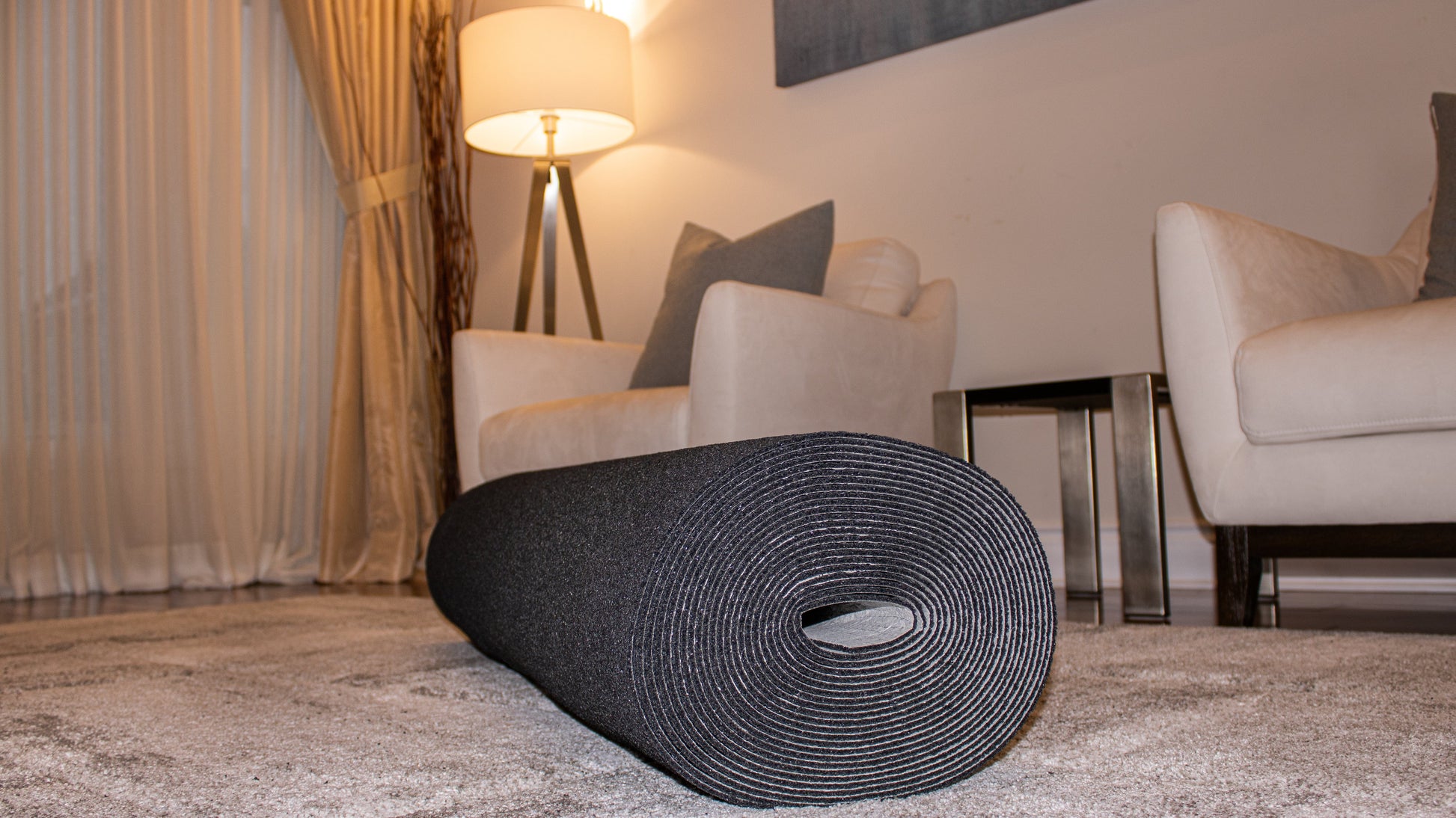 A rolled-up black yoga mat lies on a Super Dura™ 10 mm by Dura Undercushions in a living room with white armchairs, a side table, and a lit floor lamp in the background.